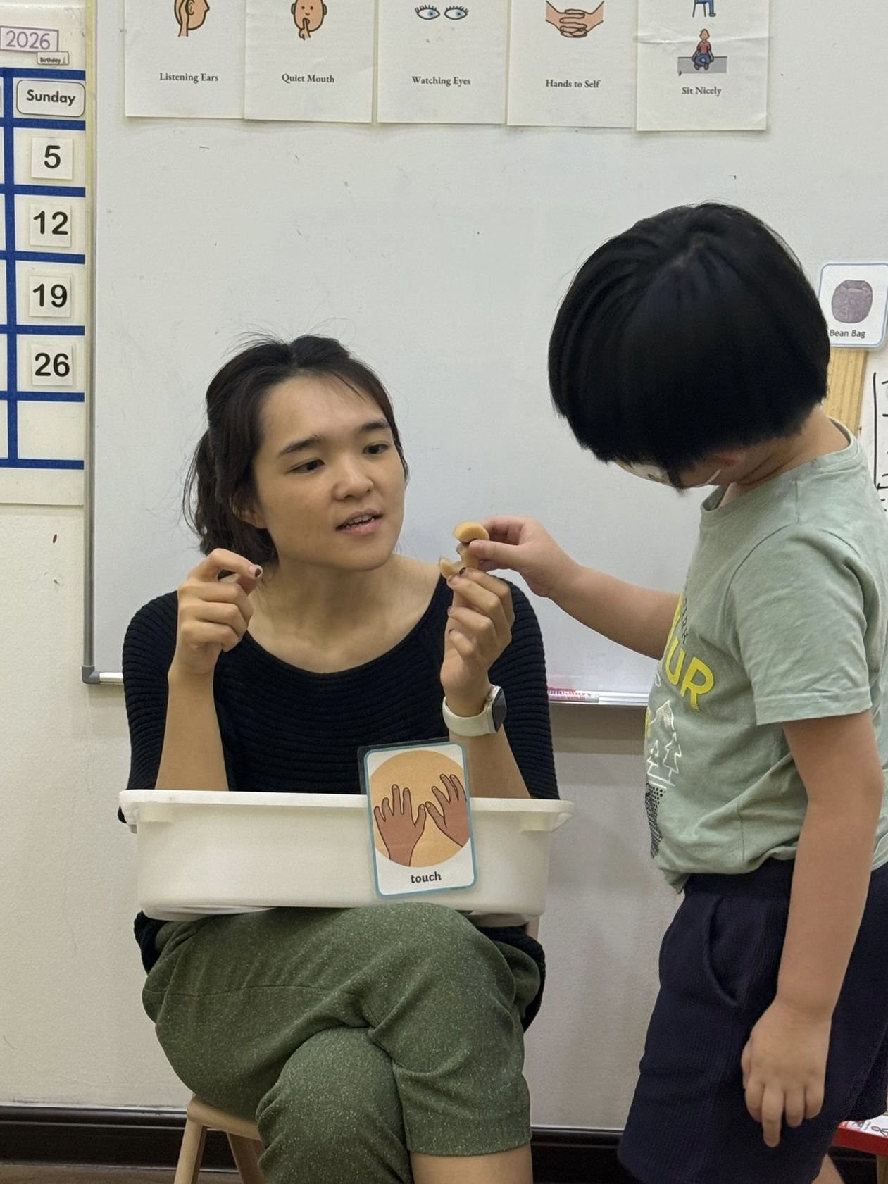 A therapist sitting with a child, holding small objects and a &lsquo;touch&rsquo; picture card, with classroom rule cards on the wall behind them